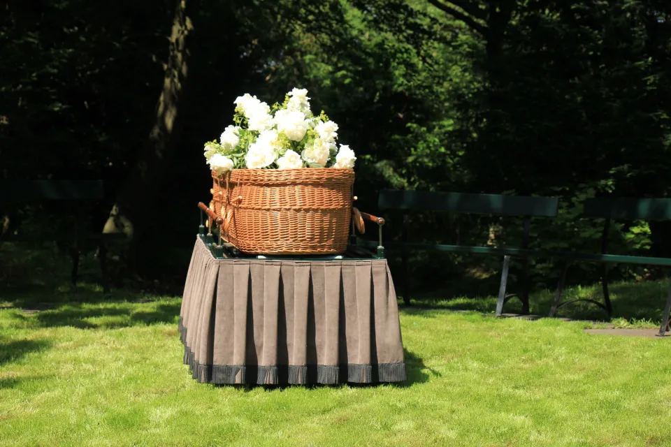 Woven willow casket with white flowers on top sits on table with pleated tablecloth on green lawn